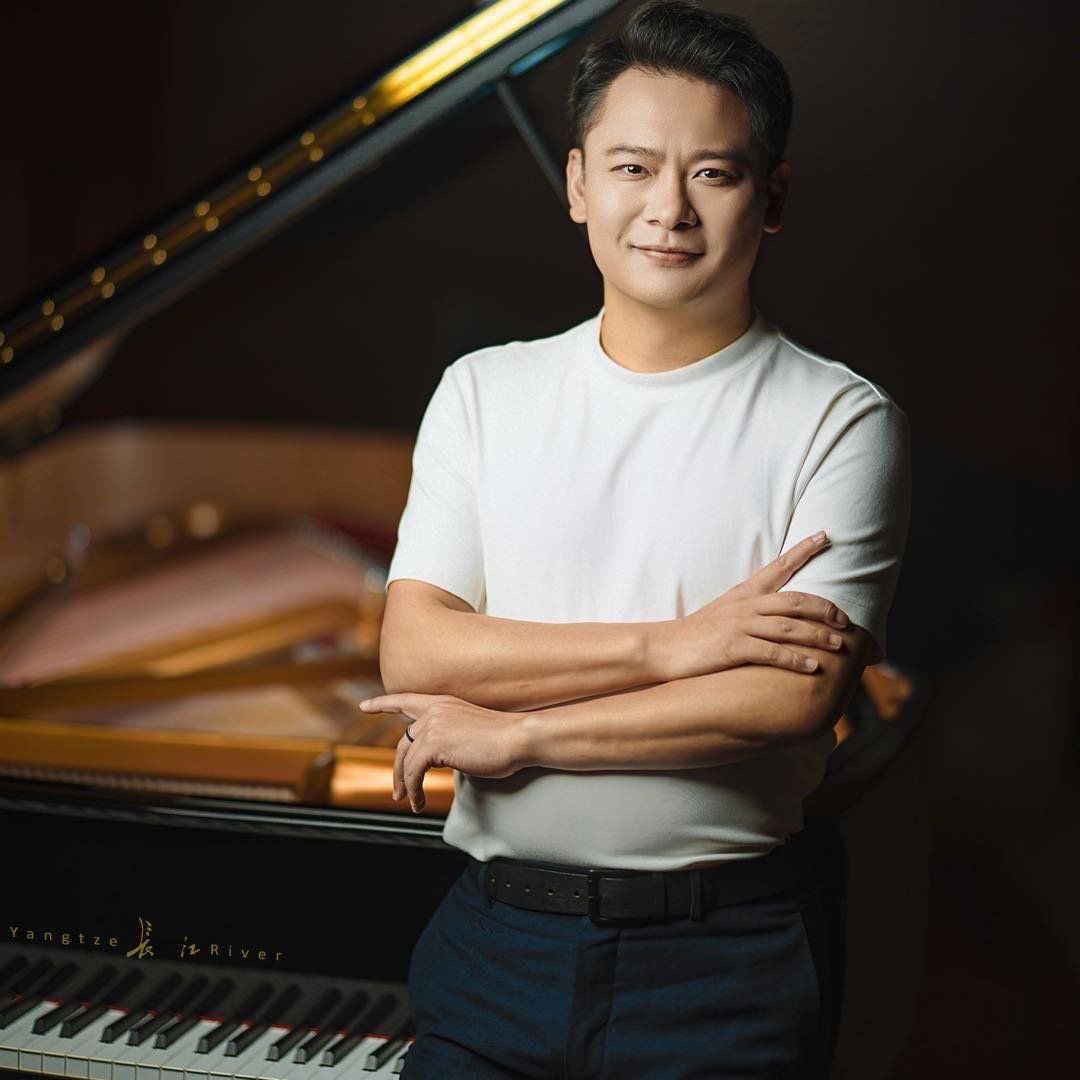 Photo of Hong Xu, a middle aged chinese man wearing a white t-shirt and dark blue dress pants, standing in front of a grand piano