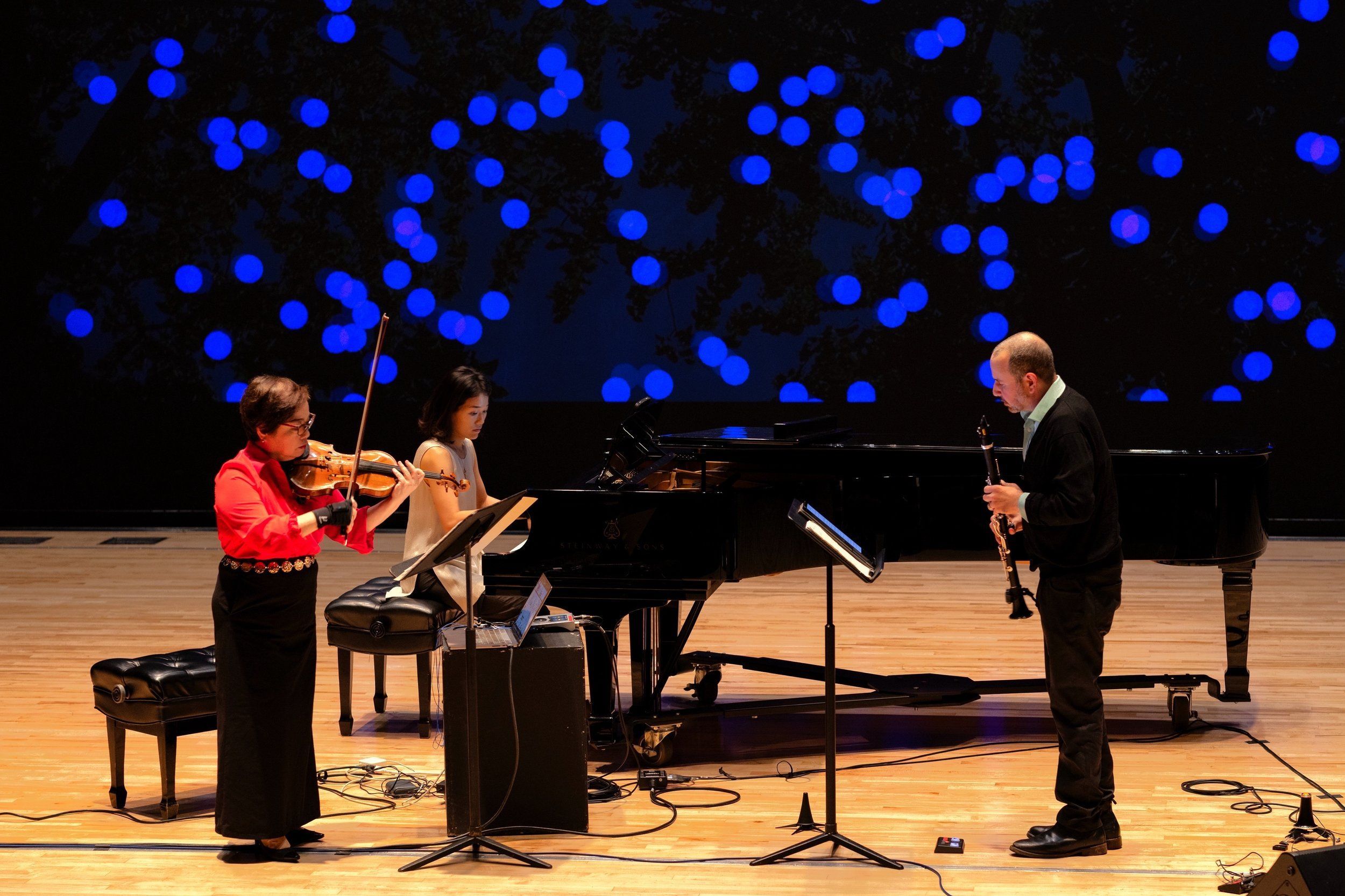 Mari Kimura, Suejin Jung and Josh Rubin playing in concert admist a blue circle animated backdrop and warm brown stage in Winifred Hall